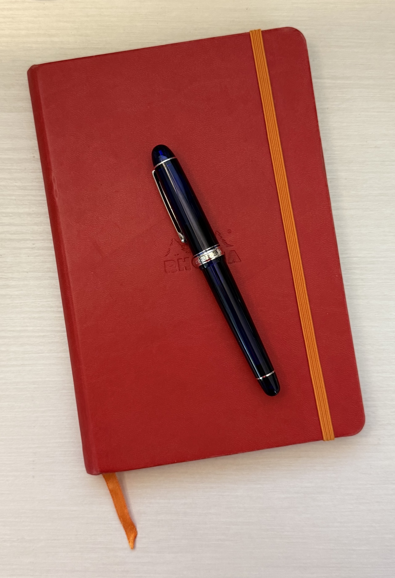 My journaling tools Platinum 3776 Century fountain pen in blue with gold accents on a red Rhodia Rhodiarama Webnotebook with an orange closure and and ribbon bookmark